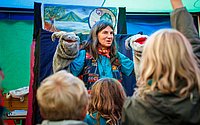  Eine Frau spricht zu Kindern, die aufmerksam zuhören. Sie steht vor einem Zelt oder Stand.