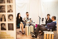 Vier Personen sitzen in einer gemütlichen Runde und nehmen einen Podcast auf. Mikrofone sind vor ihnen aufgestellt, und sie wirken vertieft in ein Gespräch. Der Raum ist hell und kreativ gestaltet, mit Bildern an einer Wand im Vordergrund.
