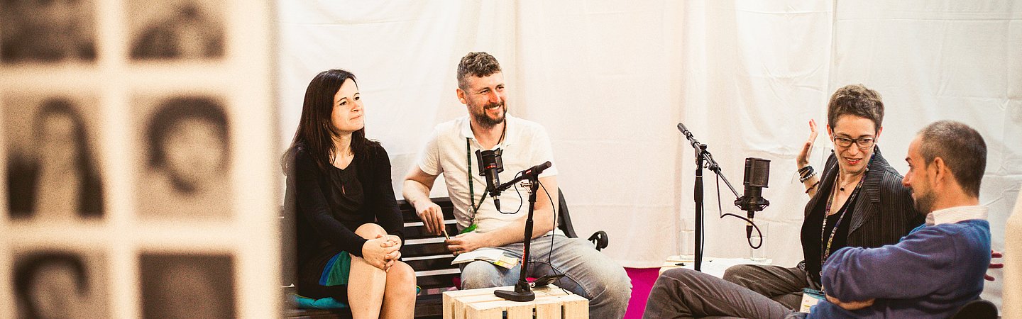Vier Personen sitzen in einer gemütlichen Runde und nehmen einen Podcast auf. Mikrofone sind vor ihnen aufgestellt, und sie wirken vertieft in ein Gespräch. Der Raum ist hell und kreativ gestaltet, mit Bildern an einer Wand im Vordergrund.