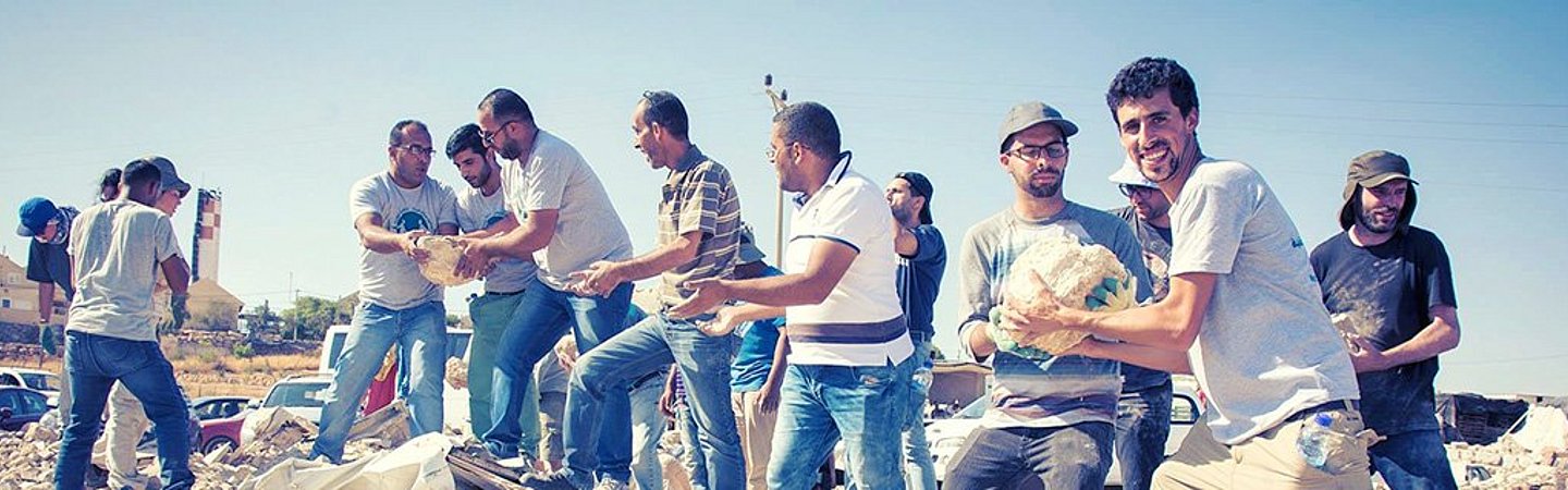Eine Gruppe von Menschen steht auf den Trümmern eines zerstörten Gebäudes. Sie tragen lässige Kleidung und blicken in die Kamera, während sie Solidarität zeigen.
