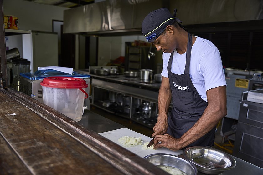 Das Bild zeigt Jailer Daniel Cortés Hortado beim Schneiden von Zutaten. Er trägt ein weißes T-Shirt und eine schwarze Schürze sowie eine schwarze Kochmütze. Im Hintergrund ist eine große, silberne Küche zu erkennen.