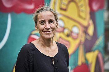 Portrait von Ulrike Purrer, einer Frau mit blonden, zurückgemachten Haaren und braunen Augen. Sie trägt eine schwarze Bluse und steht vor einer bunten Wand.