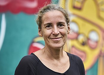 Portrait von Ulrike Purrer, einer Frau mit blonden, zurückgemachten Haaren und braunen Augen. Sie trägt eine schwarze Bluse und steht vor einer bunten Wand.