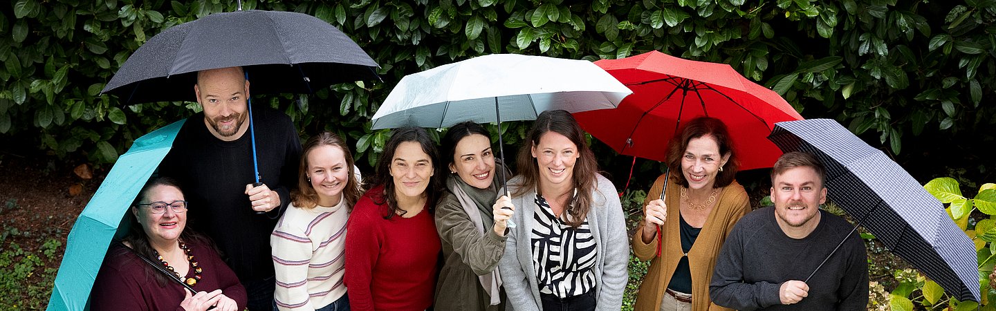 Gruppenfoto im Freien, bei dem mehrere Personen unter bunten Regenschirmen stehen. Im Hintergrund sind grüne Büsche und Blumen zu sehen. Die Gruppe lächelt in die Kamera, die Szene wirkt fröhlich und ungezwungen.
