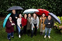 Gruppenfoto im Freien, bei dem mehrere Personen unter bunten Regenschirmen stehen. Im Hintergrund sind grüne Büsche und Blumen zu sehen. Die Gruppe lächelt in die Kamera, die Szene wirkt fröhlich und ungezwungen.