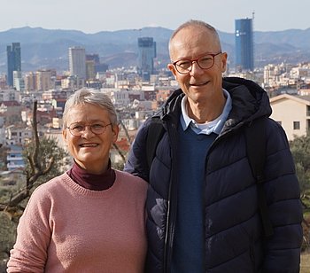 Krista und Andreas Reppich stehen nebeneinander. Im Hintergrund ist die Hauptstadt von Albanien, Tirana, abgebildet. 