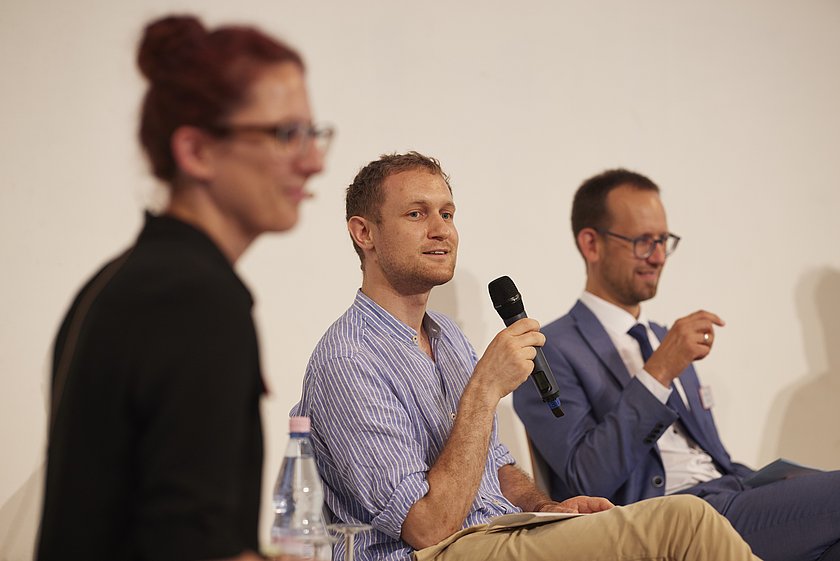 Diskussion in einem Panel, bei der mehrere Personen auf Stühlen in einem Veranstaltungsraum sitzen und miteinander sprechen.