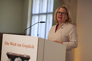 Eine Frau hält eine Rede an einem Rednerpult in einem hellen Raum.
