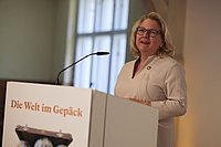 Eine Frau hält eine Rede an einem Rednerpult in einem hellen Raum.