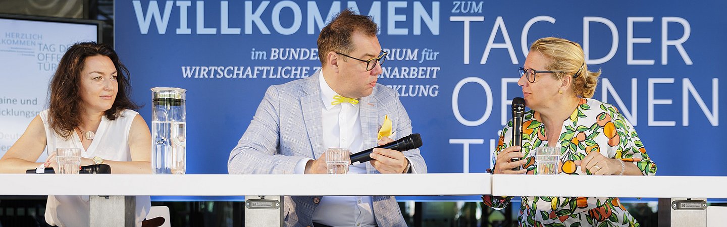 Drei Personen sitzen an einem Podiumstisch vor einer blauen Wand mit der Aufschrift 'Herzlich Willkommen - Tag der offenen Tür'. Die Szene zeigt eine Gesprächsrunde in einem offiziellen Rahmen.