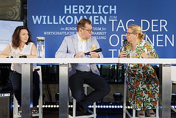 Drei Personen sitzen an einem Podiumstisch vor einer blauen Wand mit der Aufschrift 'Herzlich Willkommen - Tag der offenen Tür'. Die Szene zeigt eine Gesprächsrunde in einem offiziellen Rahmen.