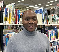 Ein Mann mit grauem Rollkragenpullover steht vor einem bunten Bücherregal.