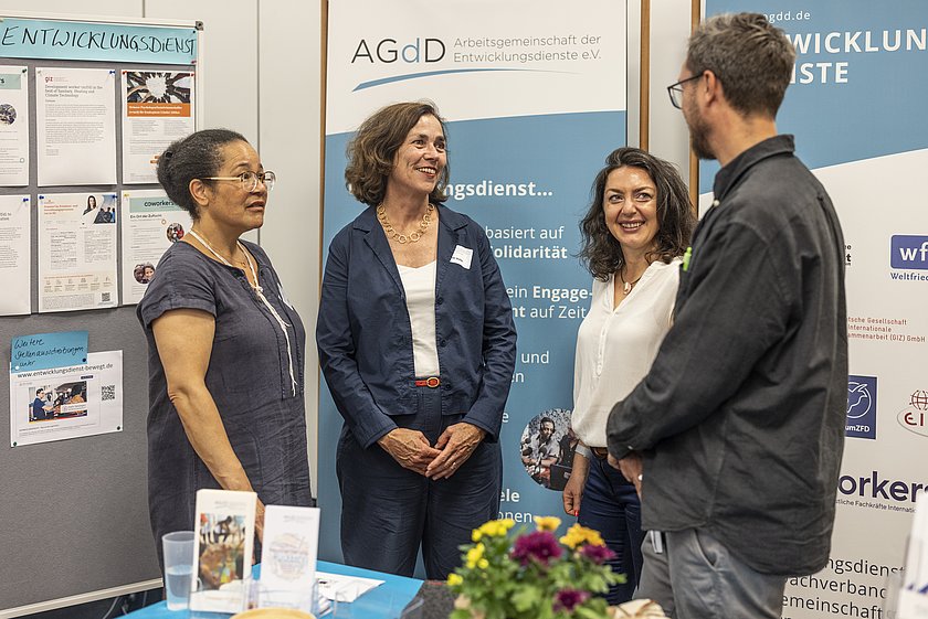 Vier Personen stehen an einem Messestand und unterhalten sich freundlich. Im Hintergrund sind Infoposter und Banner zu sehen, die den Stand dekorieren. Auf dem Tisch im Vordergrund liegen Broschüren und ein Blumenarrangement.