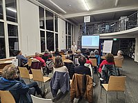 Fritz Ritzmann steht am Ende eines Raumes. Hinter ihm eine Wand mit Büchern. Neben ihm eine Leinwand. Auf der Leinwand ist eine digitale Präsentation zu sehen. Im Vordergrund sitzt das Publikum.