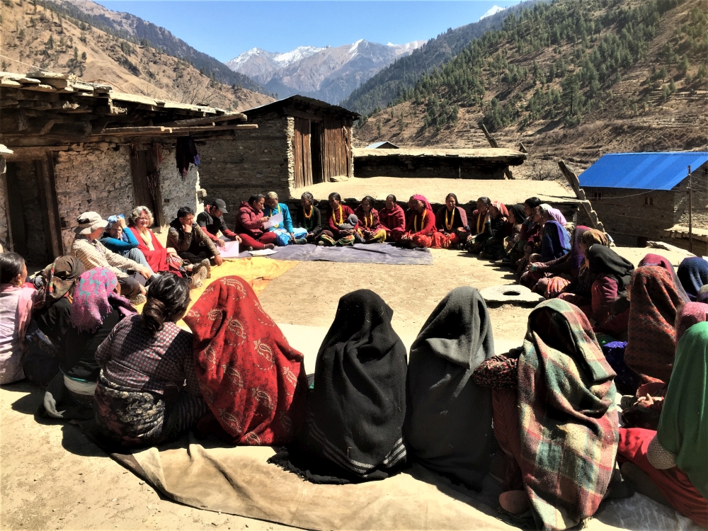 Eine Gruppe von Frauen sitzt im Freien in einem großen Kreis, viele von ihnen in farbenfrohen traditionellen Gewändern. Im Hintergrund sind einfache Steinhäuser und eine malerische Berglandschaft mit schneebedeckten Gipfeln zu sehen. Die Szene vermittelt eine gemeinschaftliche Atmosphäre in einer abgelegenen ländlichen Umgebung.